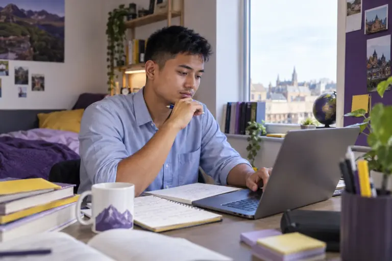 Young Nepali man with cropped hair comparing UK university courses and intake options using a laptop and notebook for UK study planning.